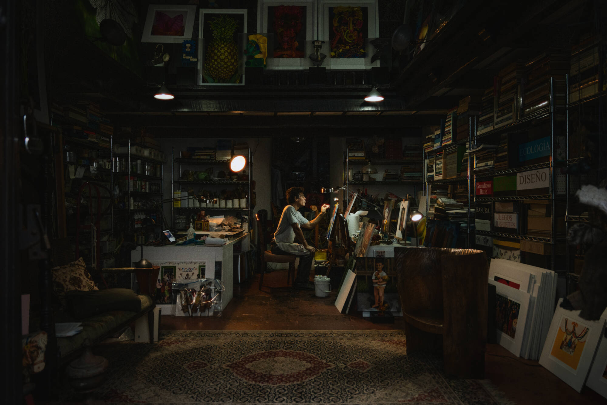A color photograph showing a person seated in profile, working on a canvas in front of them. They are sitting in a dark studio.